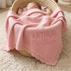 Pode incluir: Um bebê dormindo está aninhado em uma cesta de vime, coberto por um cobertor rosa de malha. O cobertor apresenta os nomes bordados "ARTHUR William" em fio rosa combinando. O cobertor tem uma borda decorativa com vieiras.