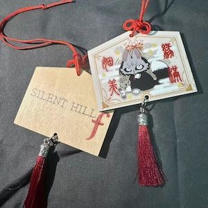 Op de afbeelding: Twee decoratieve labels met rode kwastjes en koorden. Eén label heeft de tekst "SILENT HILL" en een rode "f". Het andere label heeft een cartoonafbeelding van een grijze kat met een zwarte outfit en een kleine grijze kat. Japanse karakters zijn ook aanwezig.