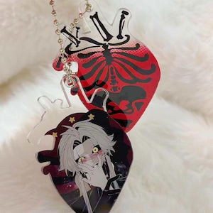 Puede incluir: Dos llaveros en forma de corazón. Uno presenta un diseño de esqueleto rojo y negro, el otro un personaje de estilo manga con cabello blanco, ojos amarillos y estrellas. Ambos están unidos a una cadena de bolas plateada.