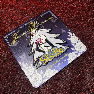 May include: A pin featuring a stylized character with long gray hair and a crown, wearing a white and black outfit with a red "R" emblem. The pin is on a dark blue card with white roses and text that reads "Jessie & Mourrow" and "Stella."