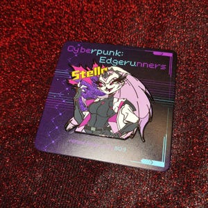 May include: A Cyberpunk: Edgerunners enamel pin featuring the character Stella. The pin has a purple, black, and pink color scheme. The pin is on a purple backing card with the text "Cyberpunk: Edgerunners" and "Stella".