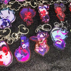 Puede incluir: Una colección de llaveros acrílicos coloridos con varios personajes de dibujos animados. Los llaveros tienen cierres y anillos de metal plateado. Los personajes están representados en tonos de rojo, morado, rosa y azul.