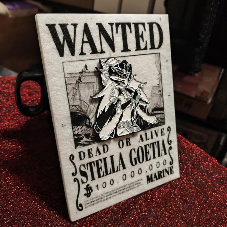 May include: A black and white enamel pin of a character with a cape and crown is affixed to a "WANTED" poster. The poster reads "DEAD OR ALIVE STELLA GOETIA 100,000,000 MARINE". The background is a black and white illustration of a ship.