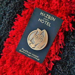 Puede incluir: Un pin de fanart de Hazbin Hotel de color cobre con un diseño de personaje, colocado sobre una tarjeta azul marino. La tarjeta dice "Hazbin Hotel" y "Pendleton copper". El pin está colocado sobre una superficie texturizada roja y negra.