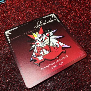 May include: A red and white enamel pin featuring a devilish anime character with the text "Sadi chan" and "Femme Fatale series NO.10 cosplay style". The pin is on a red rectangular backing.