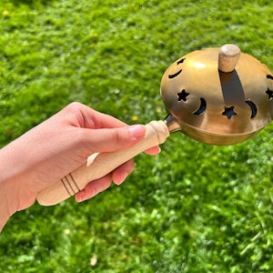 May include: A brass incense burner with a wooden handle and a lid decorated with star and crescent moon cutouts. The burner is held against a backdrop of green grass.