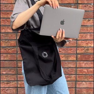May include: A person wearing blue jeans and a gray shirt is carrying a black tote bag with a white loading symbol on it. The person is holding a silver laptop computer with an Apple logo.