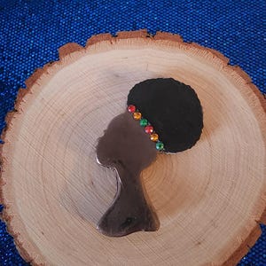 Puede incluir: Silueta marrón del perfil de una mujer con un afro negro y una diadema enjoyada de colores. La silueta está colocada sobre una rodaja de madera con vetas visibles.