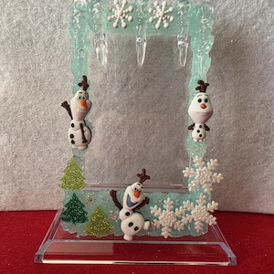 May include: A rectangular picture frame with a light blue, textured, icy border featuring hanging icicles and white snowflakes. Three Olaf figures are attached to the frame, along with three green glittery pine trees and white snowflake embellishments.