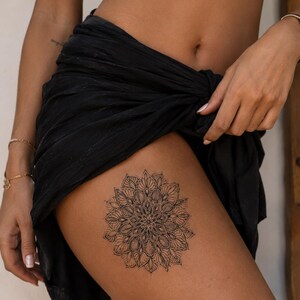 Könnte beinhalten: Ein temporäres Tattoo mit schwarzer Tinte auf dem Oberschenkel, das ein detailliertes kreisförmiges Mandala-Design mit komplexen symmetrischen Mustern, feinen Linien und blütenblattartigen Formen zeigt, die von einem zentralen Punkt ausgehen.
