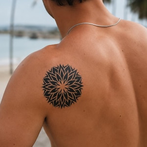 May include: A black ink tattoo on the upper back shoulder featuring a symmetrical circular mandala design with sharp, petal-like geometric patterns radiating from the centre, displayed on skin with a thin silver chain necklace visible around the neck.