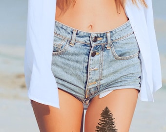 May include: A temporary tattoo featuring a detailed black ink illustration of a coniferous pine tree. The design is applied to the skin of a thigh, positioned below a pair of light blue denim shorts and a white button-down shirt worn open.