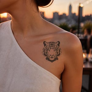 May include: A temporary tattoo featuring a detailed black ink illustration of a tiger head. The design displays the tiger's facial features, including its eyes, nose, and striped fur patterns, positioned on the upper chest area of a white garment.