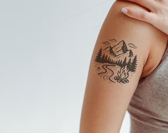 May include: A black ink temporary tattoo design applied to the upper arm, featuring a landscape illustration of a mountain range, a dense forest of pine trees, a winding river, and a small campfire with flames in a circular composition.