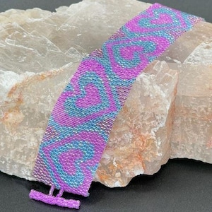 Puede incluir: Una pulsera de cuentas con un diseño de corazón morado y turquesa. La pulsera está hecha de pequeñas cuentas y tiene un cierre de barra en T. La pulsera mide aproximadamente 3,8 cm de ancho.