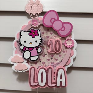 Puede incluir: Adorno para tarta rosa y blanco con temática de Hello Kitty con el nombre "LOLA" y el número "10". El adorno incluye un lazo, globos y flores decorativas. El diseño está en capas con detalles de purpurina.