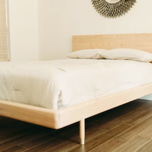 May include: A light-colored wooden bed frame with a rectangular headboard and cylindrical legs featuring metallic accents. The bed is dressed with a plain white comforter and two matching white pillows, showcasing a clean and simple design structure.