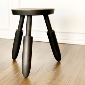 May include: A black wooden stool featuring a circular seat and three thick, tapered legs. The stool has a dark, matte finish that highlights the natural wood grain texture throughout the entire structure, including the round top and the three legs.