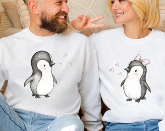 Könnte beinhalten: Zwei weiße Sweatshirts mit Pinguin-Designs. Einer der Pinguine hat eine rosa Schleife, der andere nicht. Beide haben kleine rosa Herzen. Das Paar lächelt. Die Sweatshirts haben lange Ärmel.