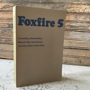 May include: A tan-colored book titled "Foxfire 5" with dark blue lettering. The book's cover lists topics including ironmaking, blacksmithing, flintlock rifles, and bear hunting. The book is standing upright.