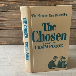 May include: Vintage hardcover book titled "The Chosen" by Chaim Potok. The cover is a light beige with blue lettering and a small illustration of three men. The spine reads "THE CHOSEN Chaim Potok".