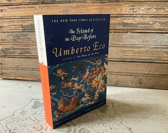 May include: A hardcover book titled "The Island of the Day Before" by Umberto Eco. The cover is navy blue with gold lettering and a celestial illustration. The spine is orange and white. The book is a New York Times Bestseller.