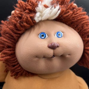 May include: A vintage doll with a round face, blue eyes, and a small nose. The doll has brown yarn hair with a white and brown accent. It is wearing a yellow-orange outfit.