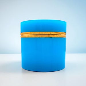 Puede incluir: Un recipiente cilíndrico azul brillante con una banda dorada en el centro. El recipiente está sobre una superficie blanca contra un fondo azul claro. El recipiente parece estar hecho de un material sólido.