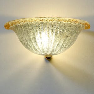 May include: A clear glass wall sconce with a textured, ribbed design. The rim is trimmed with gold, and the fixture has a gold base. The light is on, casting a warm glow on the wall.