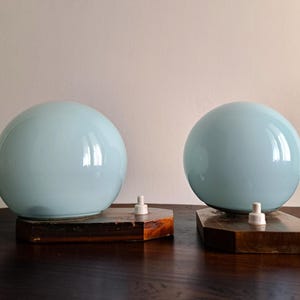 May include: Two table lamps featuring spherical, light blue glass shades mounted on dark brown, hexagonal wooden bases. Each lamp includes a small, white plastic light socket component protruding from the base next to the glass sphere.
