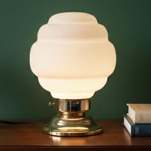 May include: A white glass table lamp with a brass base is on a dark wood cabinet. A small potted plant and two books are also on the cabinet. The lamp is lit, illuminating the room. The cabinet is approximately 100 cm wide.
