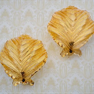 May include: Two decorative, gold-colored wall sconces shaped like stylized seashells. The sconces have a textured, ridged surface and a small, dark accent at the base. They are mounted on a cream-colored wall with a repeating floral pattern.