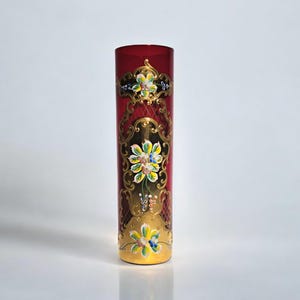 Op de afbeelding: Een rode glazen vaas met gouden en bloemmotieven. De vaas heeft een cilindrische vorm en is versierd met ingewikkelde patronen en kleurrijke bloemen.