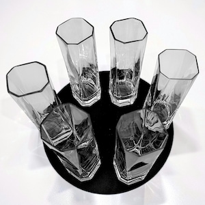 May include: Six clear, faceted drinking glasses arranged on a black circular surface. The glasses have a tall, slender shape with an octagonal rim. The image is shot from above, highlighting the glass's geometric design.