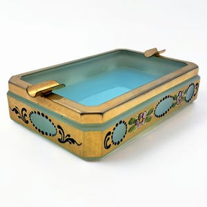 Könnte beinhalten: Rechteckiger Aschenbecher mit goldfarbenem Metallrahmen und hellblauem Innenraum. Die Seiten sind mit ovalen Designs und floralen Akzenten verziert. Der Aschenbecher hat zwei eingebaute Ablagen für Zigaretten.