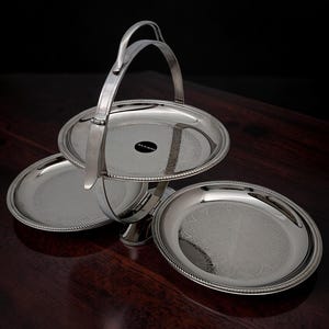 May include: A polished silver-tone metal tiered serving stand featuring three circular plates with intricate etched patterns on the surfaces. The stand includes a central vertical support column and a curved handle at the top for carrying.