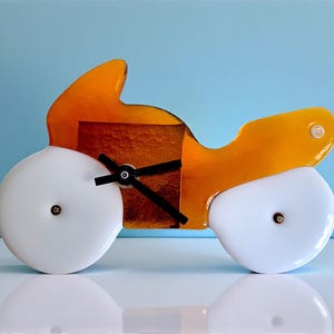 Könnte beinhalten: Eine Motorrad-förmige Uhr mit einem leuchtend orangefarbenen und braunen Körper und weißen Rädern. Die Uhr hat schwarze Zeiger und ein kleines, kreisförmiges Zifferblatt. Das Design ist modern und verspielt, vor einem hellblauen Hintergrund.