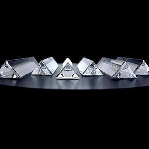May include: Six clear glass prisms arranged in a horizontal row. Each prism features a triangular cross-section with smooth, reflective surfaces that catch the light. The objects are transparent and colorless, showcasing a geometric shape.