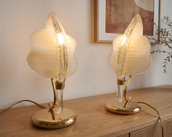 May include: A pair of table lamps featuring translucent, rippled, leaf-shaped glass shades. Each lamp has a curved, clear glass stem attached to a polished gold-tone metal base. The lamps emit a warm glow from internal light sources.