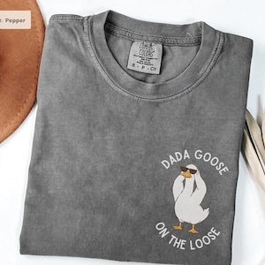 Könnte beinhalten: Ein graues T-Shirt mit dem Text "DADA GOOSE ON THE LOOSE" und einer Cartoon-Gans mit Sonnenbrille. Das Shirt hat einen Rundhalsausschnitt und ein Comfort Colors-Etikett.