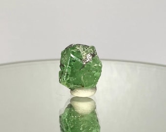 May include: A vibrant green crystal with a rough, textured surface. The gemstone is displayed on a small, white, rounded base, with its reflection visible below. The crystal's color is a bright, saturated green, with hints of darker shades.