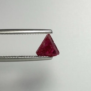 May include: A small, triangular-shaped deep red gemstone held by a pair of silver-coloured metal tweezers. The stone features a faceted surface with visible texture and a vibrant, saturated crimson hue, contrasting against the metallic tool.