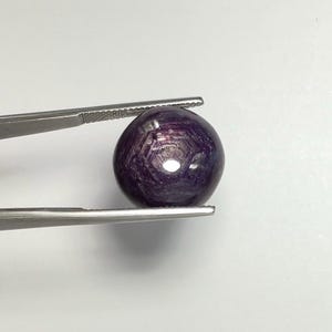 May include: A spherical, deep purple gemstone is held by silver tweezers. The gem has a polished surface with internal patterns and a reflective sheen. The tweezers are made of stainless steel.