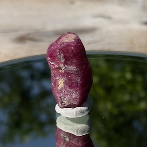 May include: A raw, deep magenta-colored ruby crystal with a rough, textured surface and irregular facets. The stone has a cylindrical, elongated shape with a slightly rounded top and sits upright on a small, light-colored circular base.