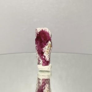 May include: A raw, elongated gemstone with a deep magenta hue, speckled with white and beige inclusions. The mineral specimen is displayed on a reflective surface, showcasing its natural, unpolished texture.