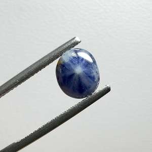 May include: A polished, oval-shaped blue gemstone with a star-like pattern. The gem is held by a pair of silver tweezers against a white background. The gem's surface reflects light, showcasing its unique internal structure.