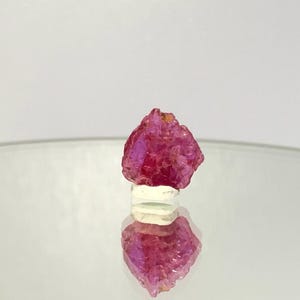 May include: A rough ruby gemstone in a vivid pink-red shade, set on a small, white base. The image shows the gemstone's reflection below. The background is a soft, neutral white.