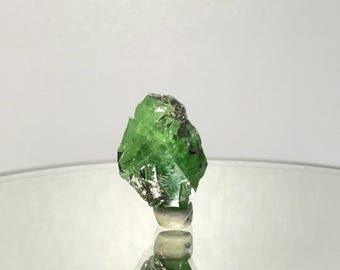 May include: A raw, uncut green gemstone with dark inclusions. The gem is displayed on a reflective surface, showcasing its natural, irregular shape and vibrant color. The gemstone is a mineral specimen.