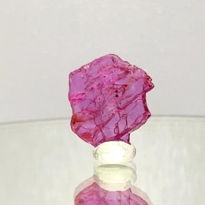 May include: A rough-cut, pink gemstone with a crystalline structure. The gem is mounted on a white base, reflecting light. The background is a soft, neutral colour, highlighting the gem's vibrant hue. This is a natural mineral specimen.