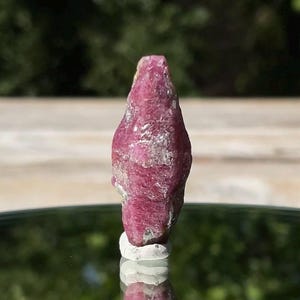 May include: A raw, unpolished ruby crystal with a deep magenta-pink hue and a naturally irregular, elongated hexagonal shape. The surface of the gemstone displays a textured, crystalline structure with visible facets and opaque mineral inclusions.